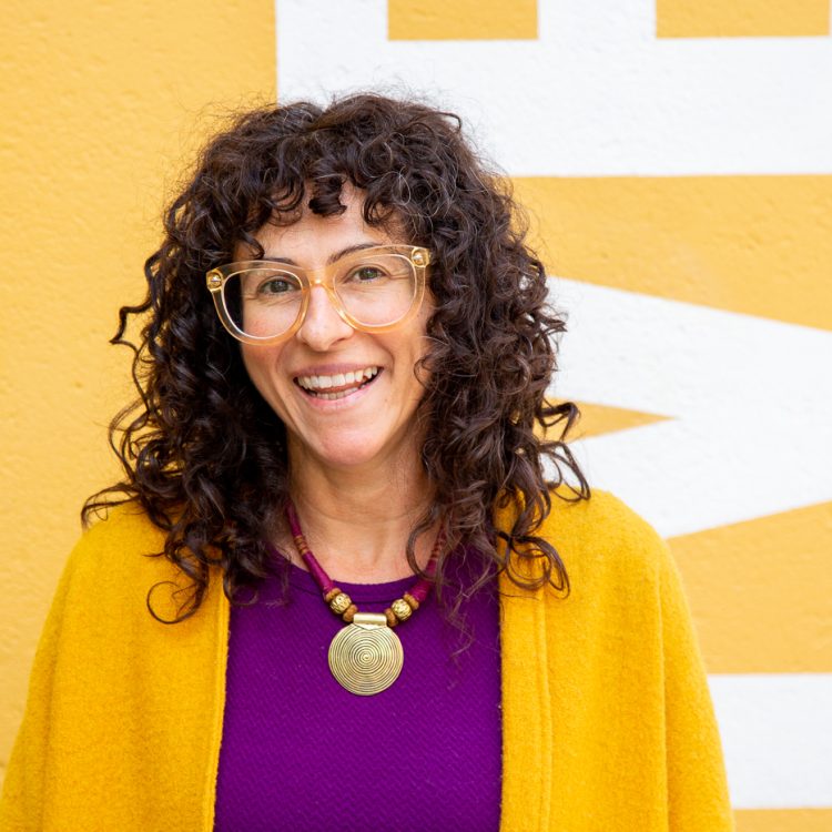 A photo of a woman in a yellow shall and purple shirt with a yellow wall behind her